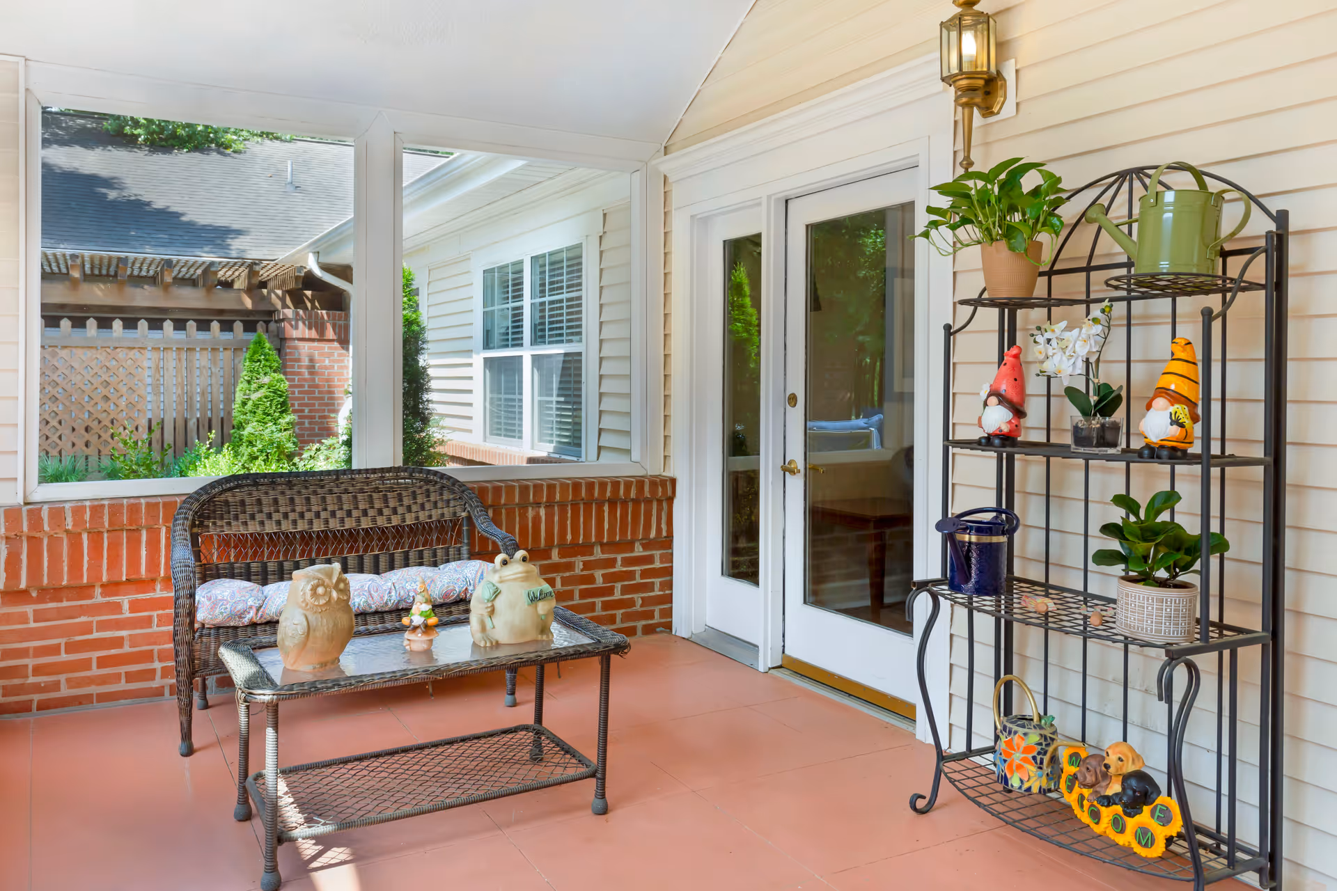 A screened-in porch area with a wicker bench and glass-top table displaying decorative owl and frog figurines. To the right, a black metal shelving unit holds various potted plants and garden gnome decorations. The porch has brick half-walls, a tiled floor, and white double doors leading inside.