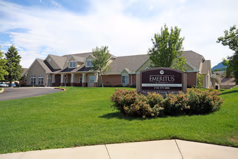 Exterior view of a senior living facility building with a well-maintained green lawn and a sign in front that reads 'Emeritus at Broadmoor' with contact information. The building has a beige and brick facade with multiple windows and a sloped roof under a partly cloudy sky.