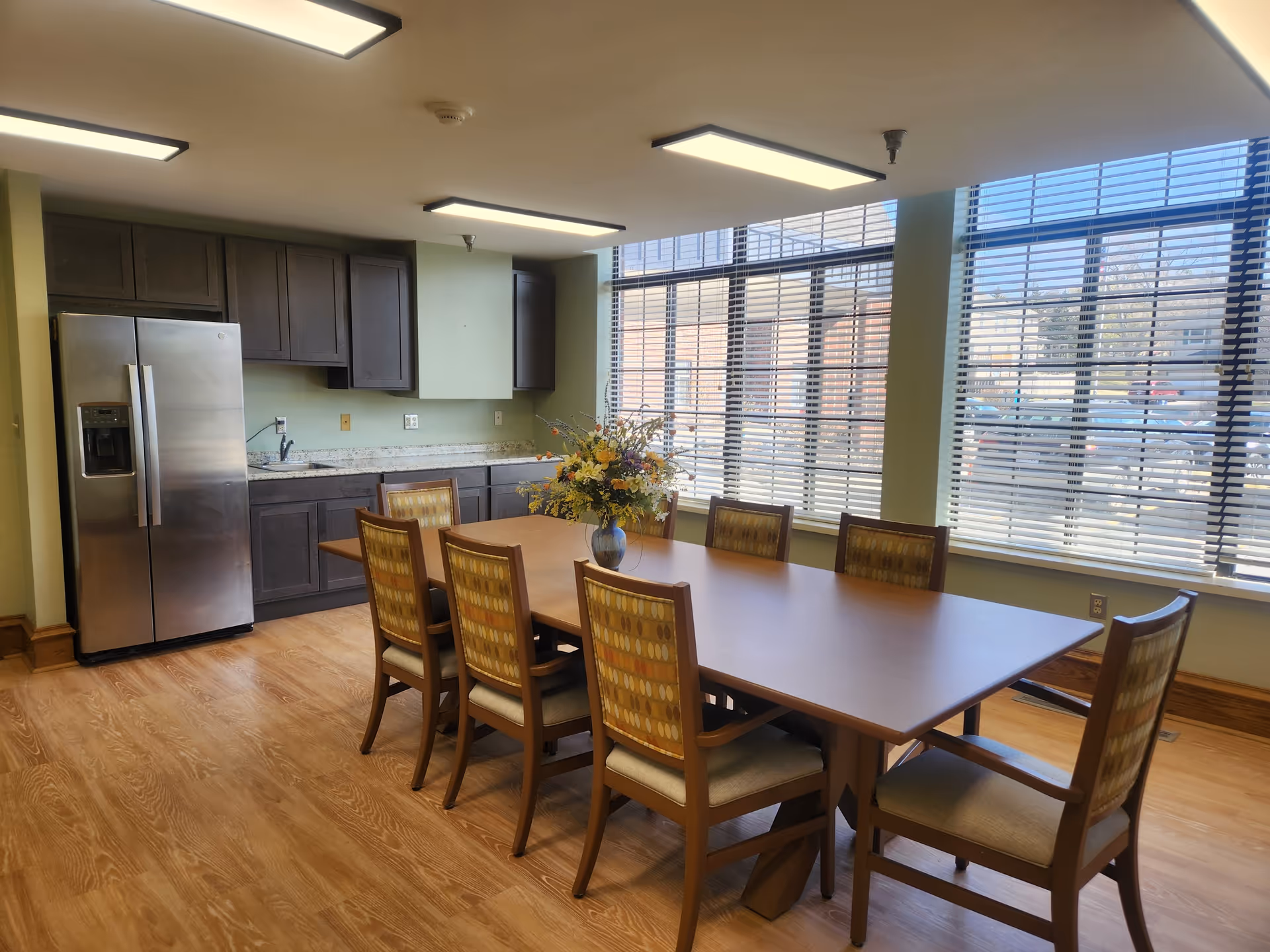 A dining area with a long wooden table surrounded by eight cushioned chairs with patterned backs. A vase with a colorful flower arrangement is placed in the center of the table. Behind the table is a kitchenette with dark cabinets, a stainless steel refrigerator, a sink, and a granite countertop. Large windows with horizontal blinds allow natural light to fill the room, and the floor is covered with wood-patterned flooring.