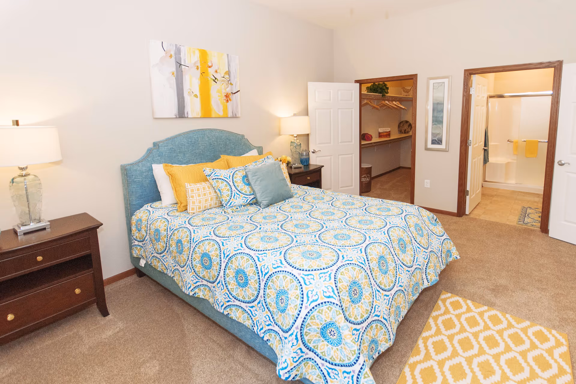 A bright bedroom with a blue upholstered bed featuring a colorful patterned bedspread and multiple pillows. There are two wooden nightstands with lamps on either side of the bed. The room has beige carpet and light-colored walls with a piece of abstract art above the bed. An open door reveals a walk-in closet with shelves and hanging space, and another door leads to a bathroom with a shower and yellow towels.