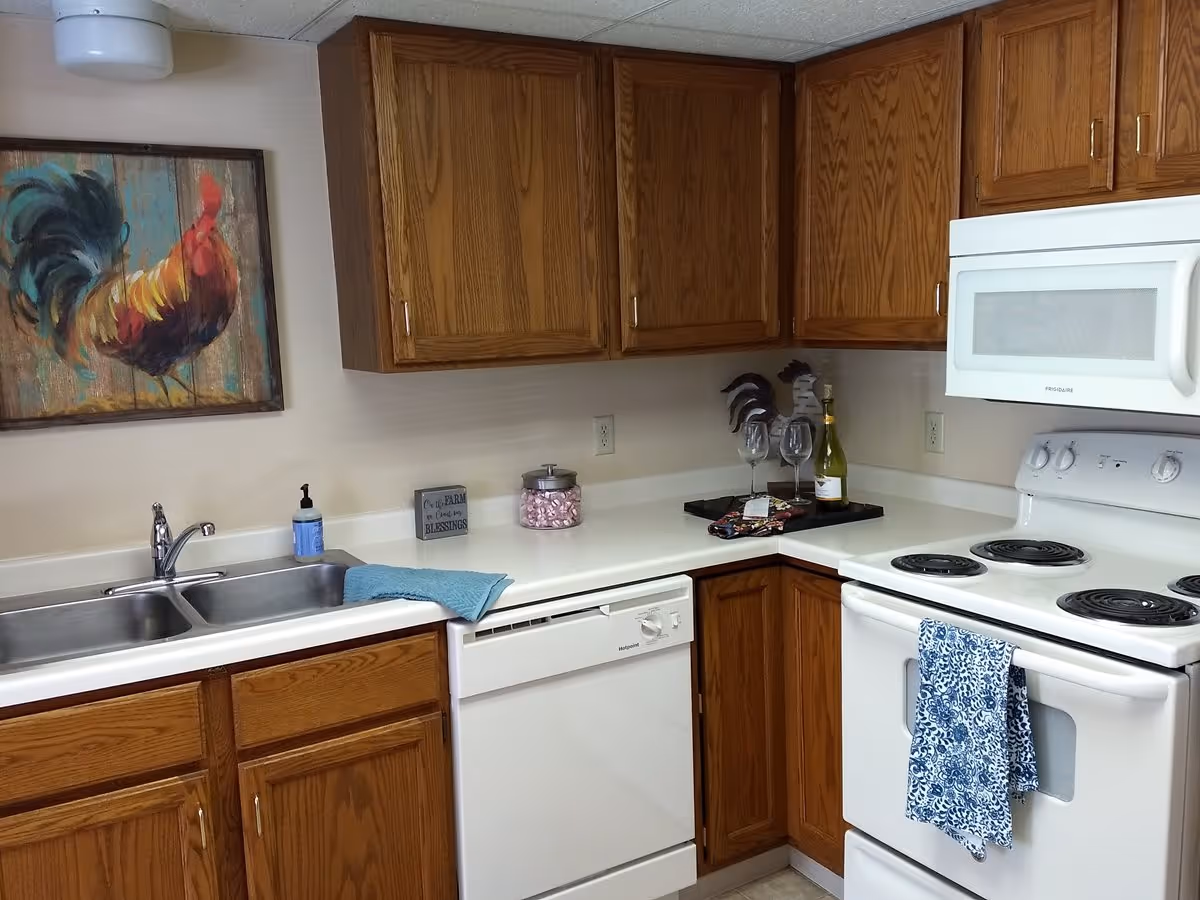 A kitchen corner with wooden cabinets, a white electric stove with a blue patterned towel hanging on the handle, a white microwave above the stove, a white dishwasher, and a double sink with a blue cloth draped over the edge. On the countertop, there is a soap dispenser, a small decorative sign, a jar filled with pink wrapped candies, two wine glasses, a bottle of white wine, and a tray with a napkin and some small items. A colorful painting of a rooster hangs on the wall above the sink.