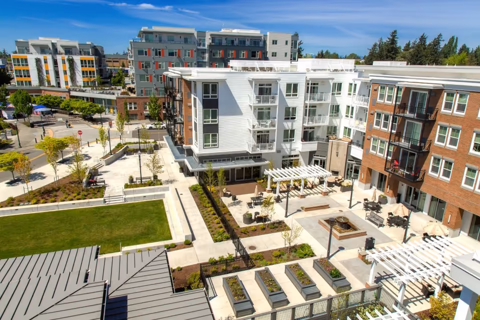 Aerial view of a modern senior living complex courtyard with multi-story buildings, patios, seating areas, landscaping, and walkways under a blue sky.