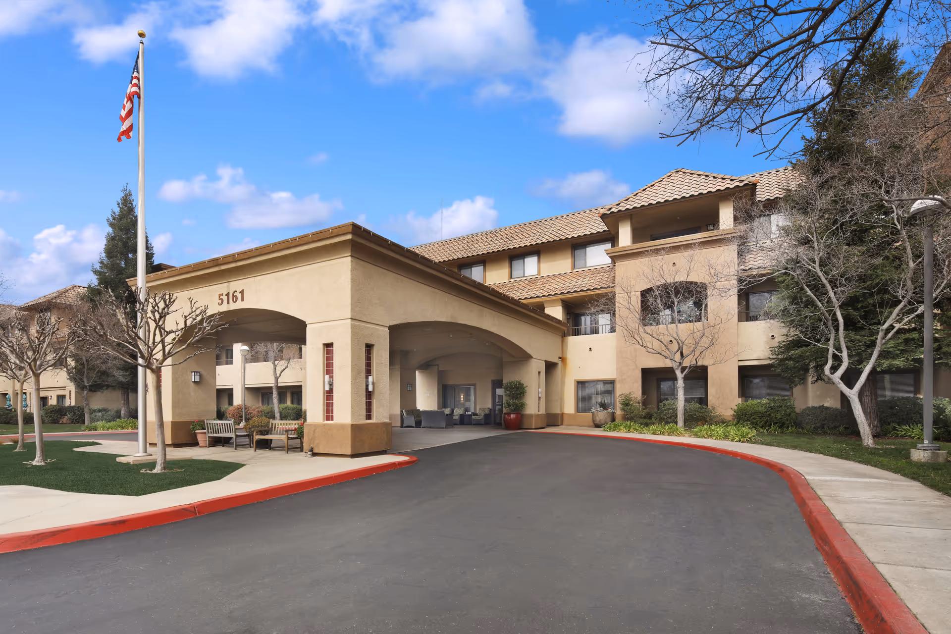 Exterior view of a senior living facility building with beige walls and a tiled roof. The entrance has a covered drop-off area with the number 5161 displayed. There are leafless trees, benches, a flagpole with an American flag, and a curved driveway with a red curb. The sky is partly cloudy.