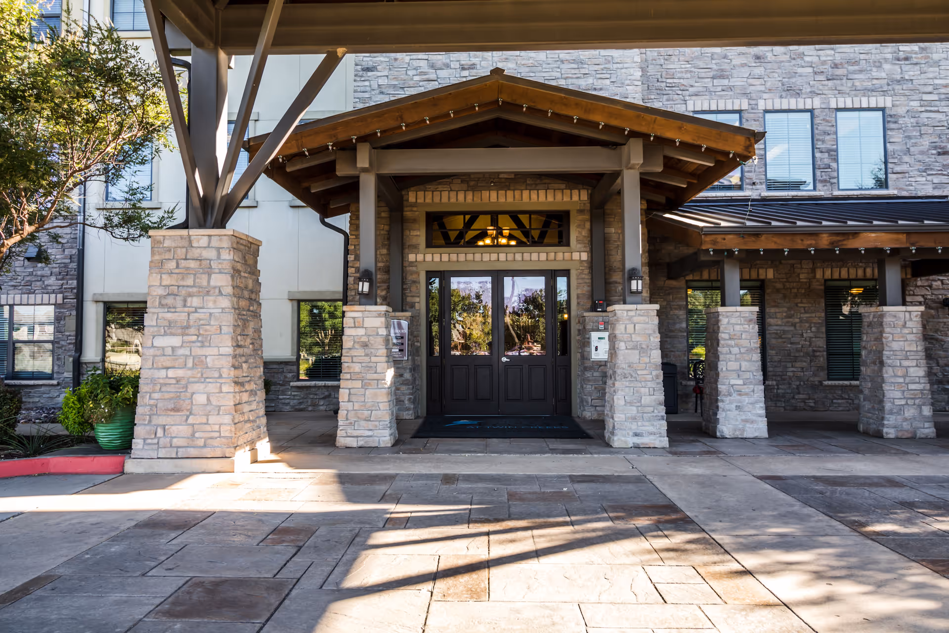 Front entrance of a building with a covered wooden portico, stone pillars, and double glass doors.