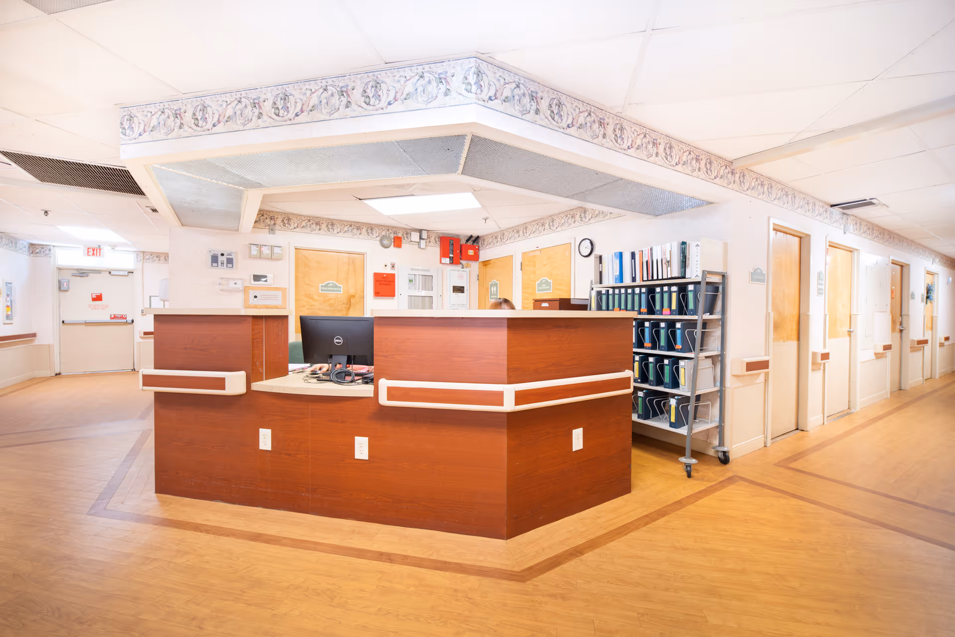A central nurse station/reception desk in a bright hallway of a senior living facility with a computer, file cart, and doors to resident rooms.