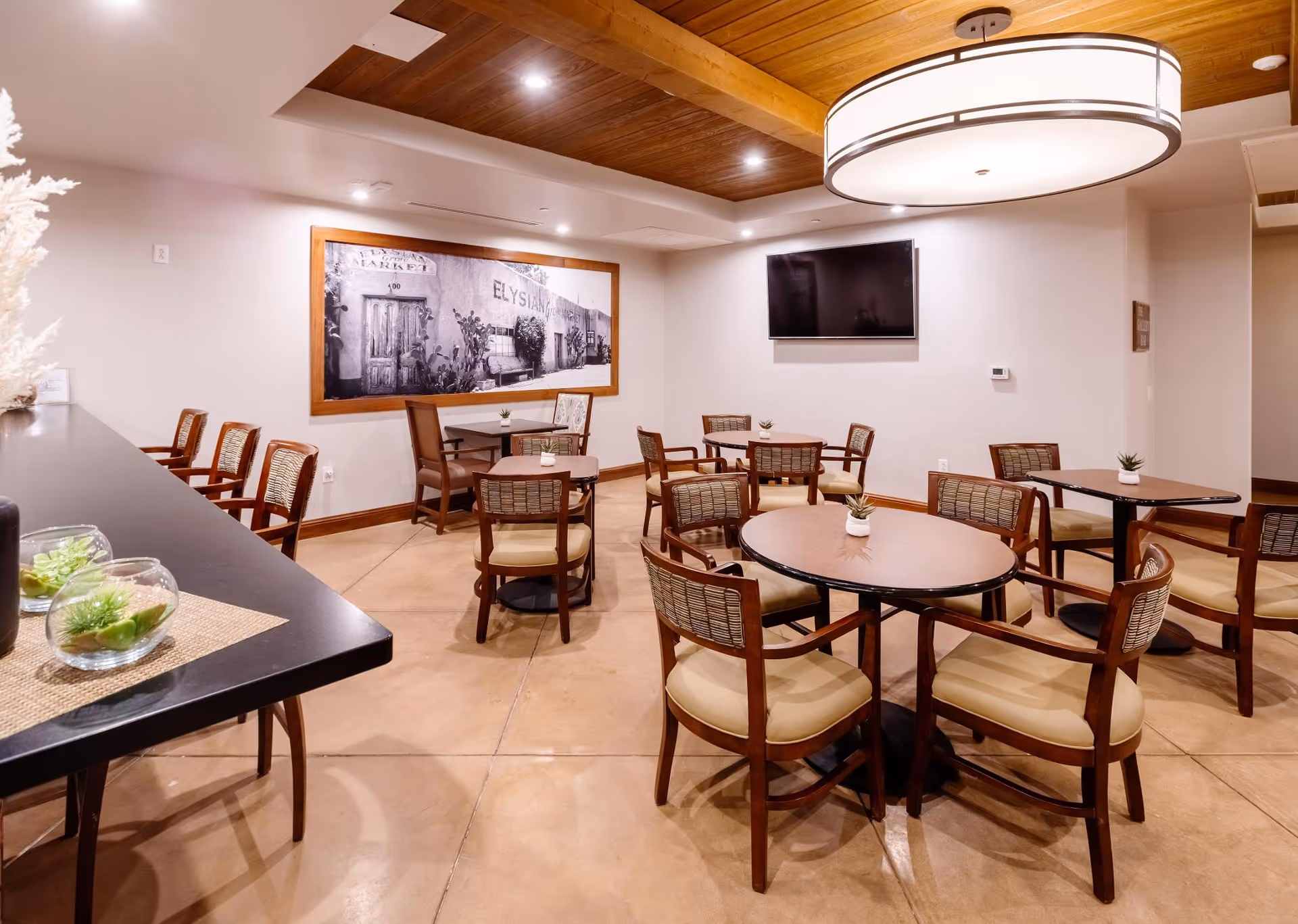 Interior view of a dining area in a senior living facility with multiple round and square tables surrounded by wooden chairs with cushioned seats. A large modern light fixture hangs from a wooden ceiling section. A flat-screen TV is mounted on the wall, and a large framed black and white photograph decorates another wall. Small potted plants are placed on the tables and counter.