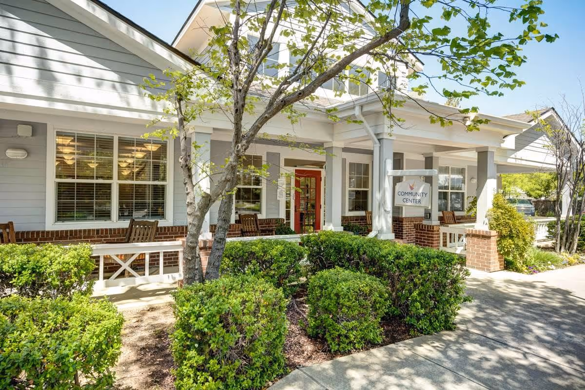 Exterior view of a community center building at Ivy Park at Laguna Creek with a porch, wooden benches, green bushes, and a tree in the foreground under a clear blue sky.