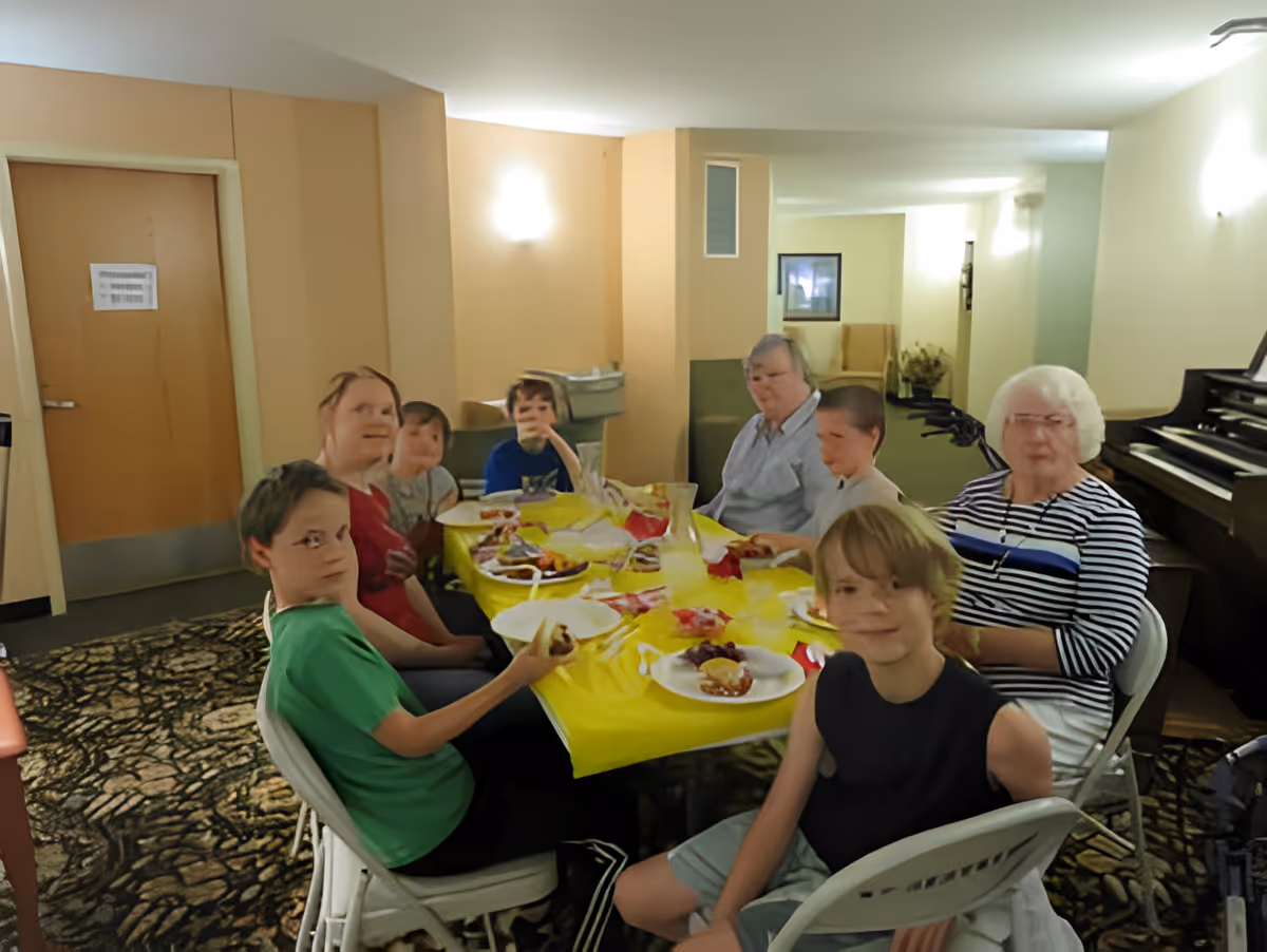 A group of people, including children and elderly individuals, sitting around a table covered with a yellow tablecloth, eating and socializing in a well-lit room with beige walls and a patterned carpet. There is a piano visible on the right side of the room.