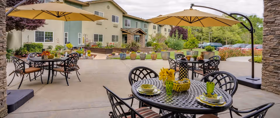 Outdoor patio with metal dining tables, umbrellas, and potted plants in front of a multi‑story residential building.