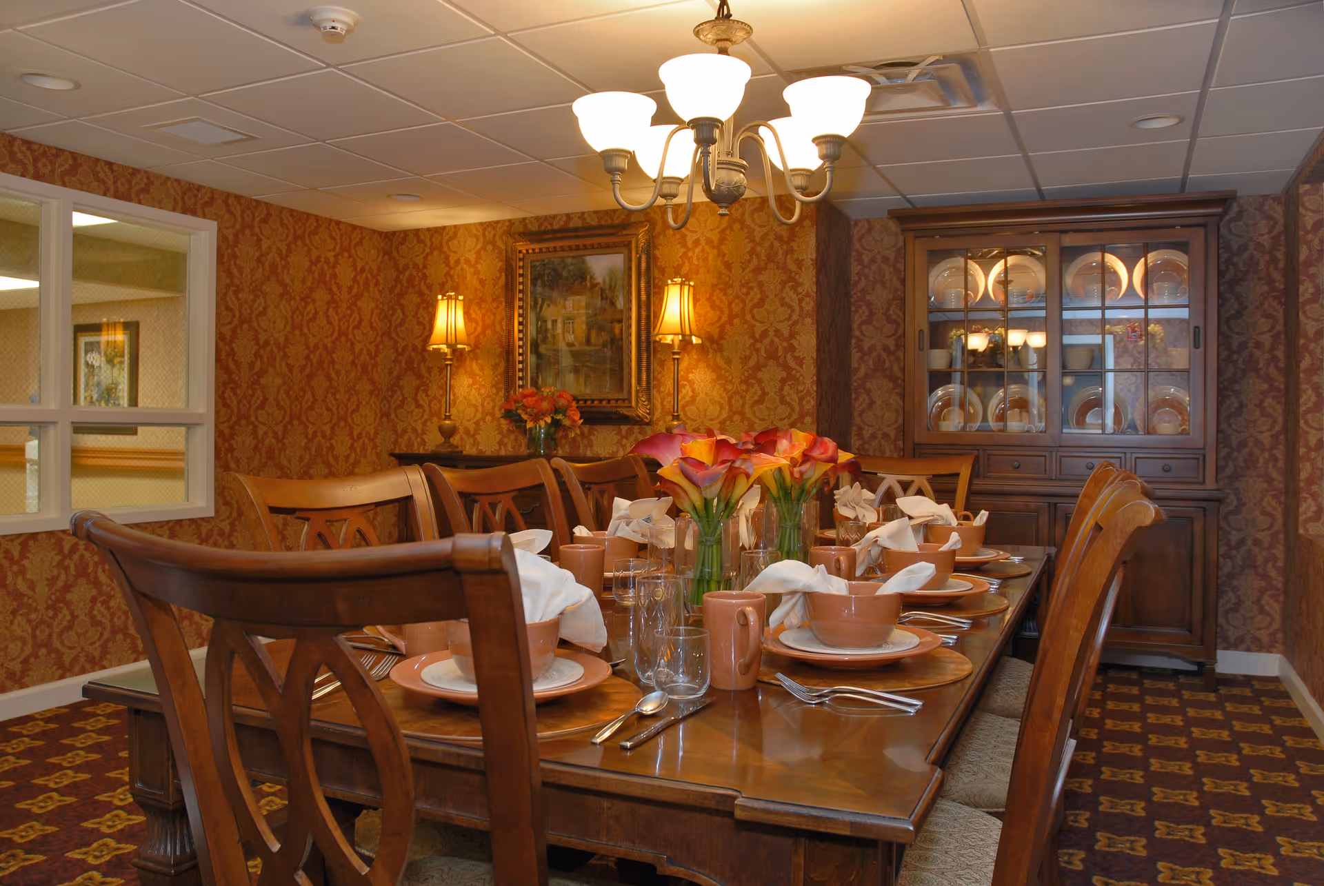 A warmly lit dining room with a long wooden table set for a meal, featuring brown dishware, glasses, silverware, and white napkins. The table is decorated with vases of orange and yellow flowers. The room has patterned wallpaper, a chandelier overhead, two wall lamps, a framed painting, and a wooden china cabinet filled with plates.
