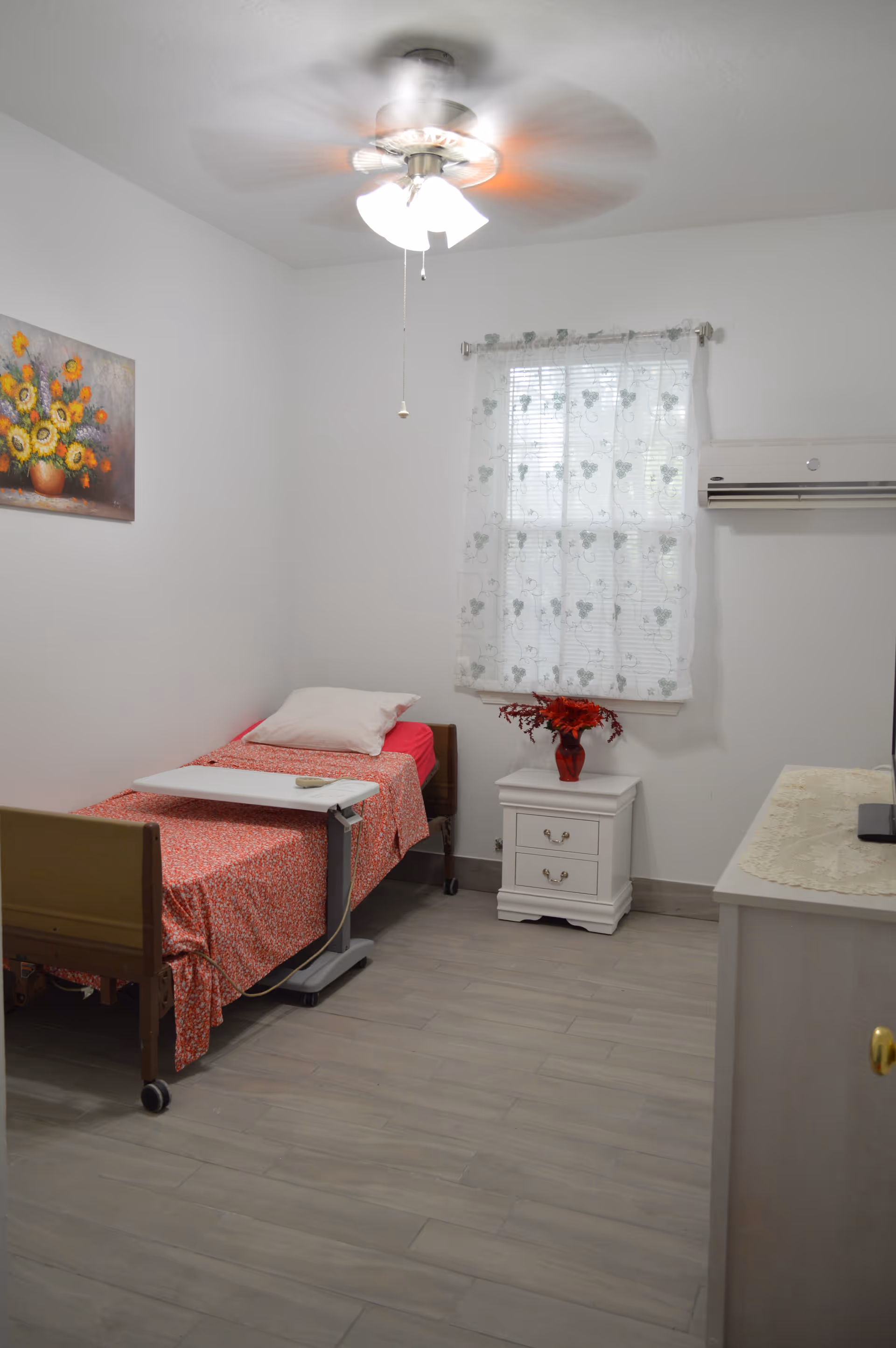 A small, simple bedroom with a single bed covered in a red patterned blanket and a white pillow. There is a white overbed table on the bed. A white nightstand with two drawers is next to the bed, holding a red vase with red flowers. A window with sheer white curtains featuring a floral pattern is on the wall above the nightstand. A ceiling fan with lights is on the ceiling. A painting of sunflowers hangs on the wall to the left. The floor is light-colored wood, and a dresser with a lace runner is partially visible on the right side.