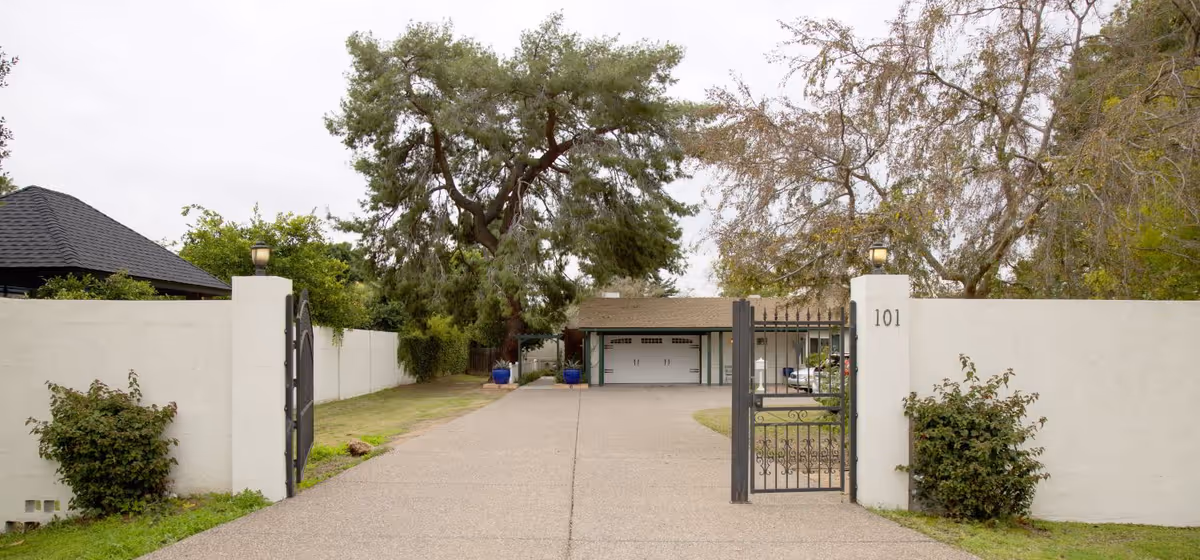 Entrance to Tangelo Grove Senior Care Home featuring a wide driveway leading to a building with a garage door, flanked by white walls and black metal gates, with trees and shrubs on both sides.