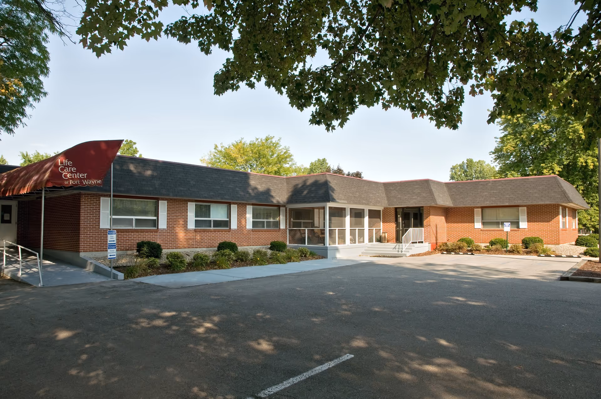Exterior view of a single-story brick building with a dark shingled roof and a red awning that reads 'Life Care Center of Fort Wayne'. The building is surrounded by trees and landscaping, with a paved parking area in front.