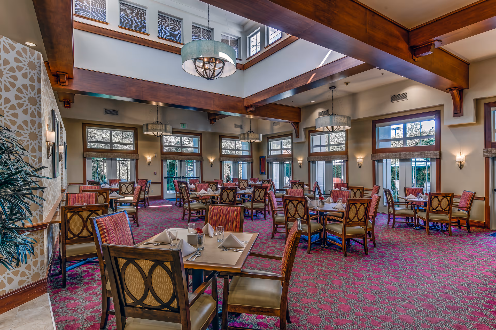 Bright, spacious dining room with multiple set tables and chairs, large windows, and exposed wooden ceiling beams.