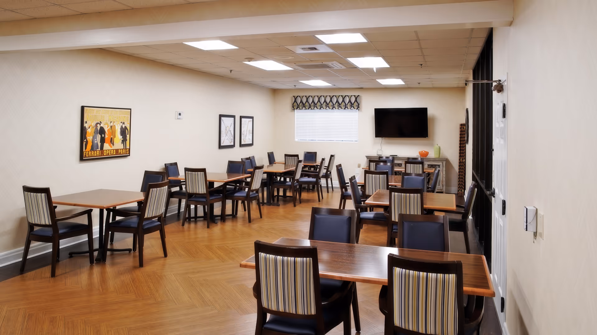 Dining/activity room with multiple wooden tables and striped-upholstered chairs, hardwood floor, wall-mounted TV and framed artwork.