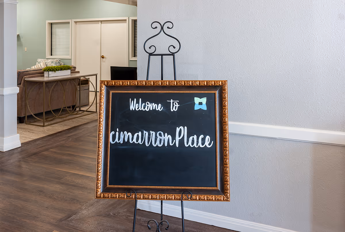 An interior view of a senior living facility entrance area with a decorative framed chalkboard sign on an easel that reads 'Welcome to cimarronPlace'. In the background, there is a seating area with a couch, a table with a plant, and double doors.