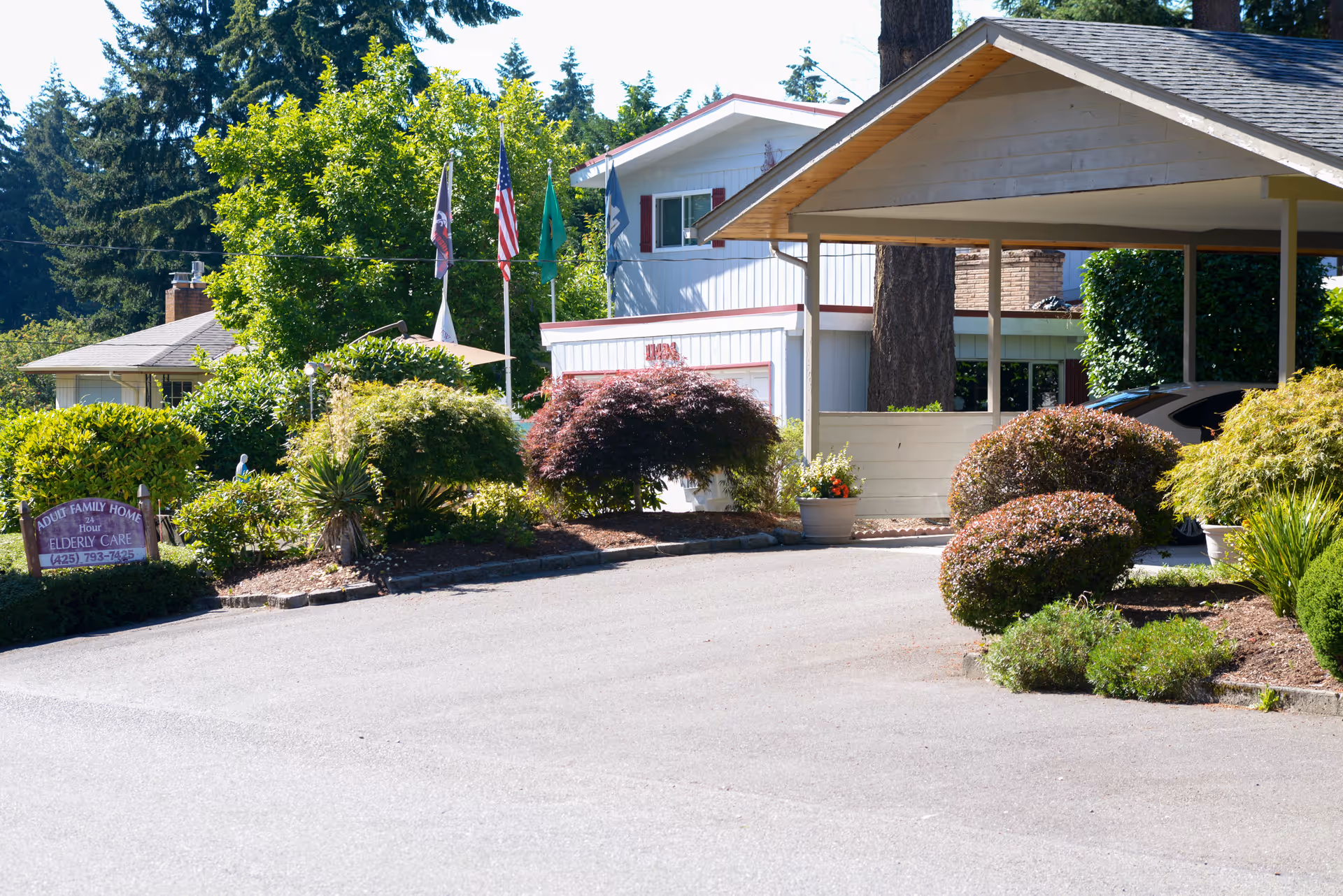 Driveway and landscaped front entrance of a senior living home with a carport, flags, and a small sign.