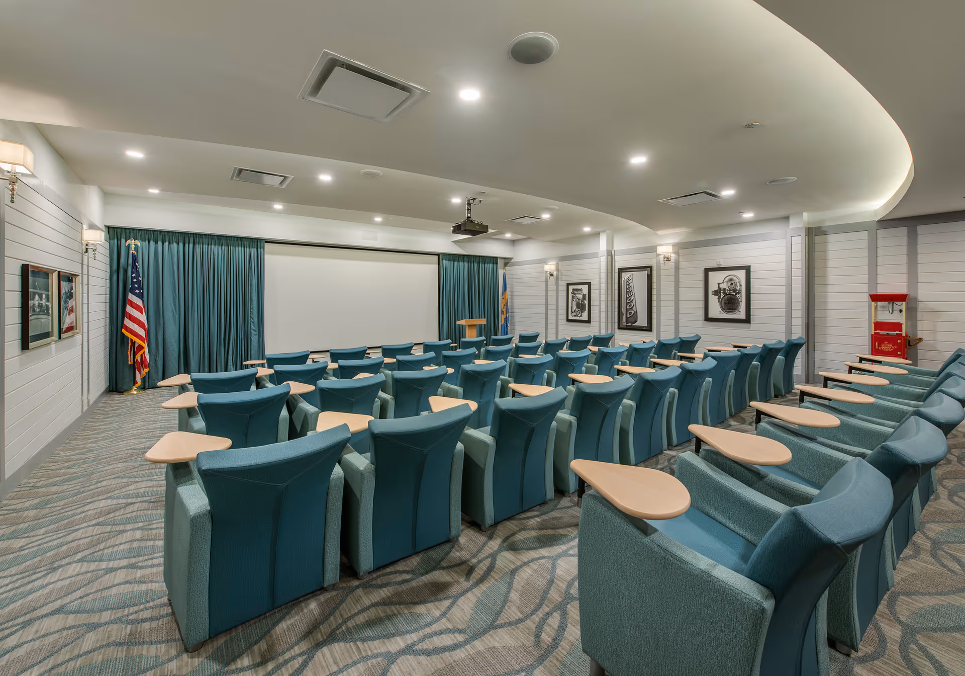 A conference or presentation room with rows of teal upholstered chairs, each equipped with a small wooden writing surface. At the front of the room is a large white projection screen flanked by teal curtains, an American flag, and a wooden podium. The walls are decorated with framed black and white photographs, and the ceiling features recessed lighting and a curved design.