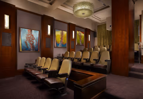 A small theater or screening room with two rows of comfortable beige and brown reclining seats. The room features wood-paneled walls with colorful abstract paintings and soft wall lighting. A large decorative ceiling light fixture hangs above, and heavy curtains cover the windows on the right side.