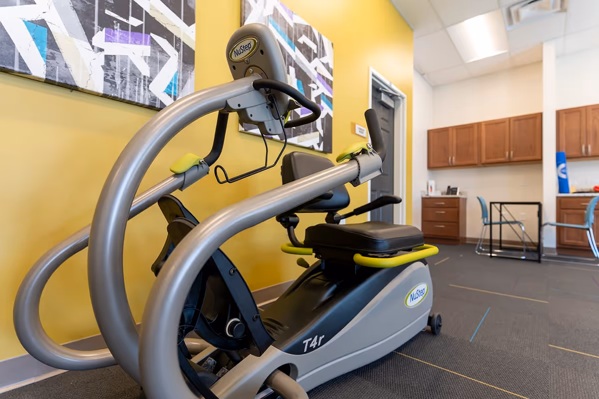 A NuStep recumbent exercise machine in a bright fitness/rehab room with yellow walls and wooden cabinets.