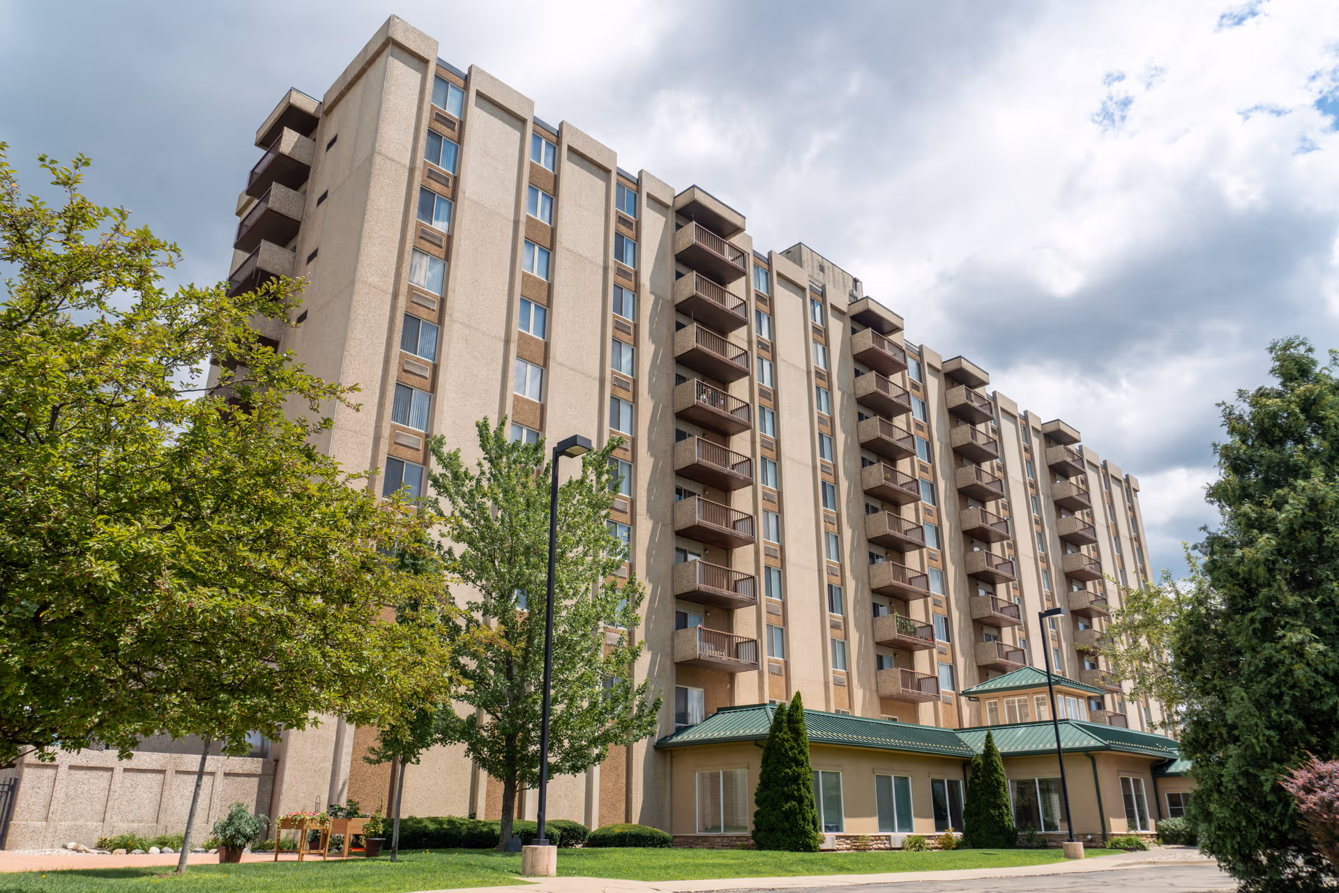 Exterior view of a multi-story senior living facility building with balconies, surrounded by trees and greenery under a partly cloudy sky.