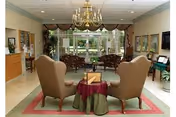 Lobby/lounge area with two wingback chairs around a small table, additional seating, a chandelier, and large windows overlooking greenery.