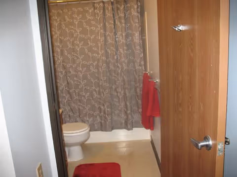 Open door reveals a bathroom with a toilet, a shower behind a patterned curtain, and red towels and a matching bath mat.