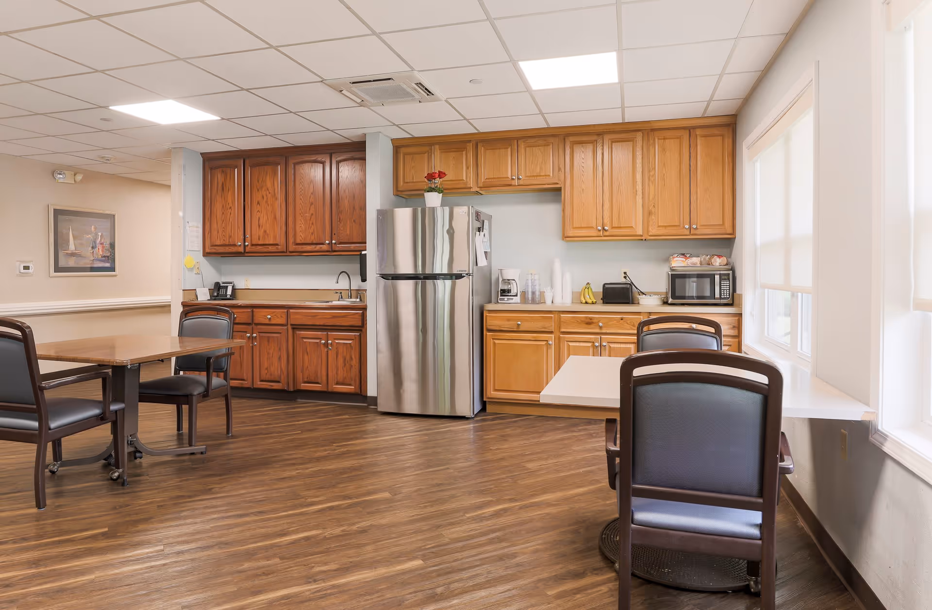 A well-lit kitchen and dining area in an assisted living facility featuring wooden cabinets, a stainless steel refrigerator, a microwave, a coffee maker, and tables with chairs. The room has wood flooring and large windows with blinds allowing natural light to enter.