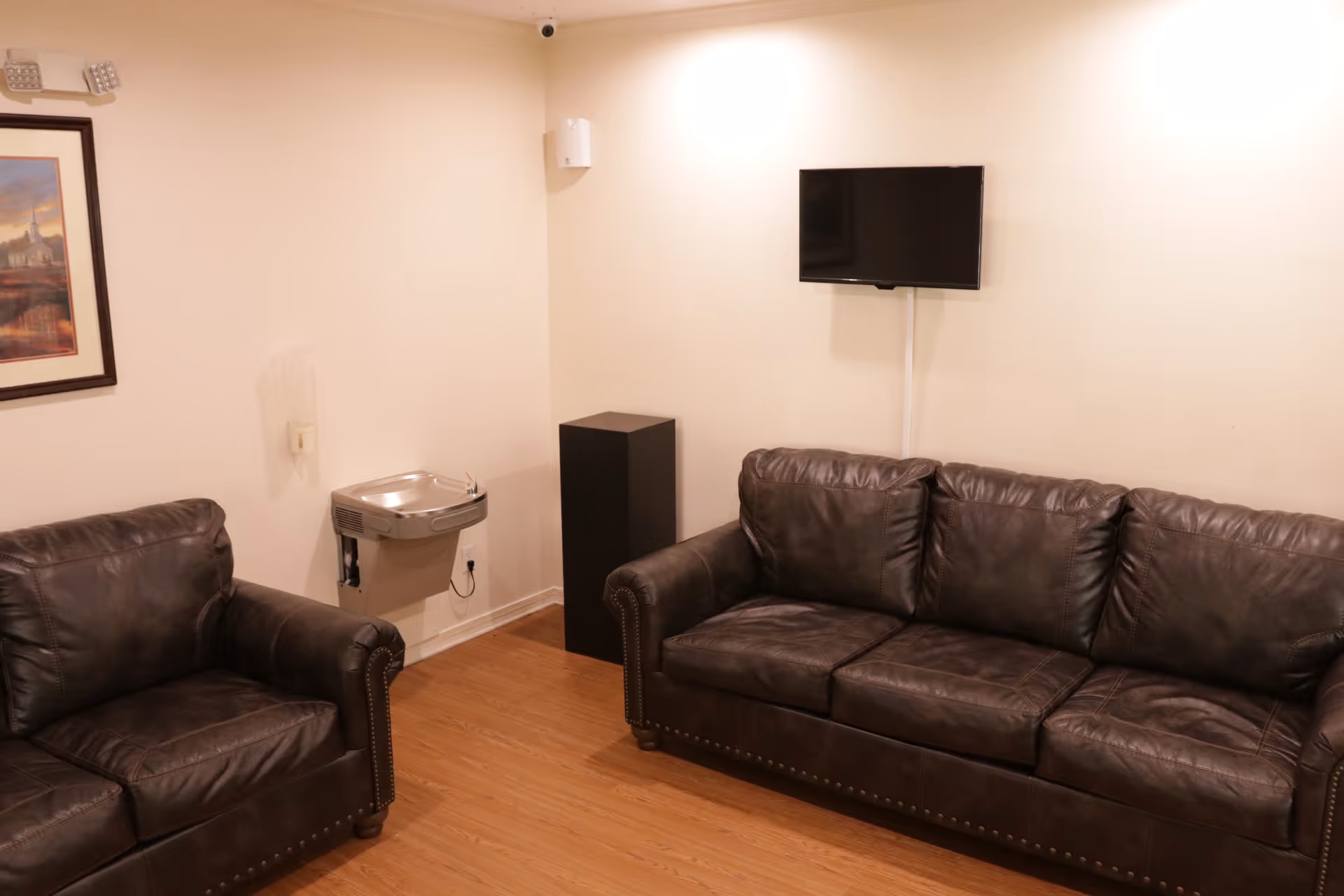 A small lounge area with two dark brown leather sofas facing each other, a wall-mounted flat screen TV, a water fountain, a black speaker or cabinet, and a framed picture on the wall. The room has light-colored walls and wooden flooring.