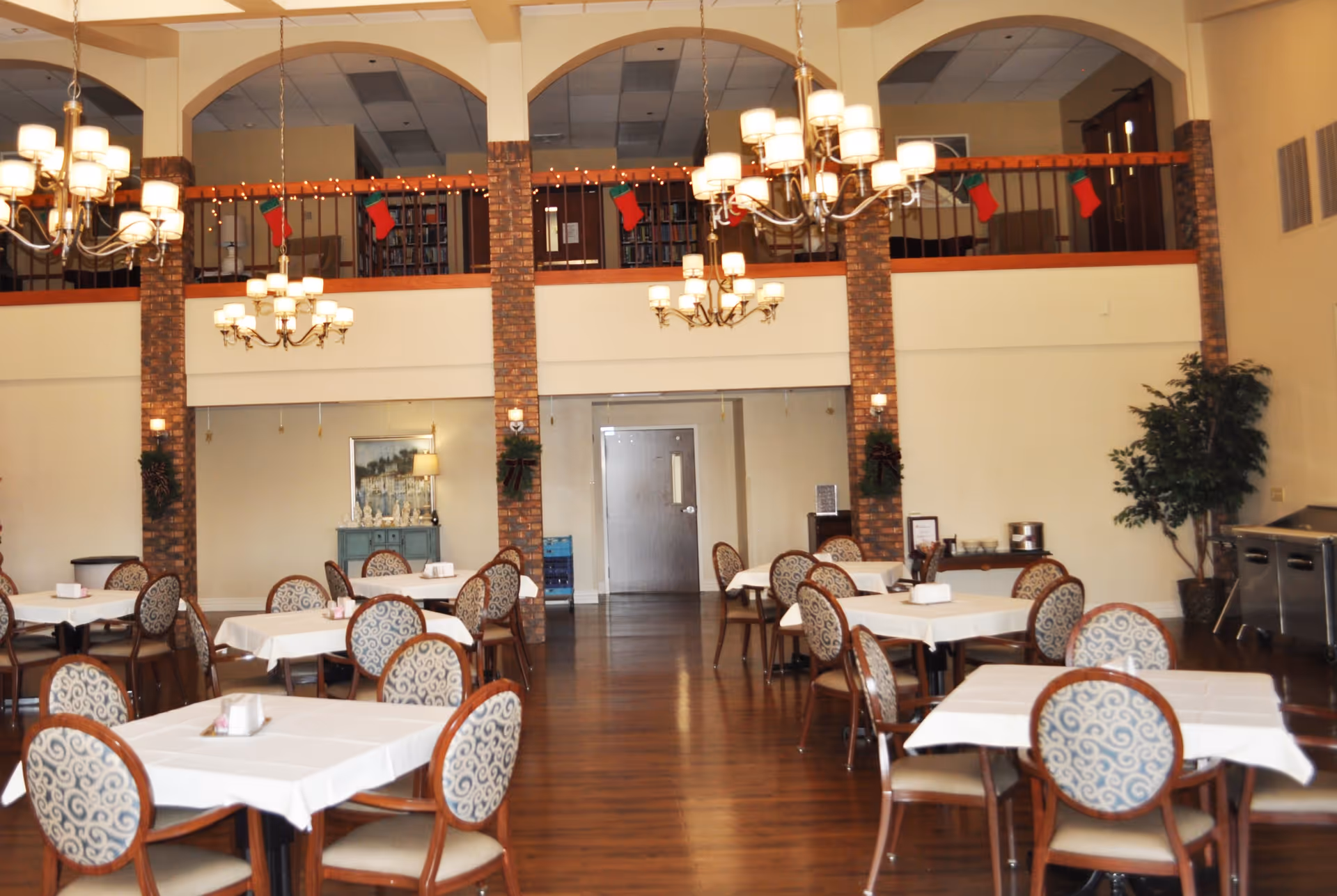 A spacious dining room with multiple tables covered in white tablecloths and surrounded by patterned chairs. The room features high ceilings with chandeliers, brick pillars, and a balcony decorated with Christmas stockings. There is a door at the far end, a small cabinet with a lamp, and a potted plant on the right side.