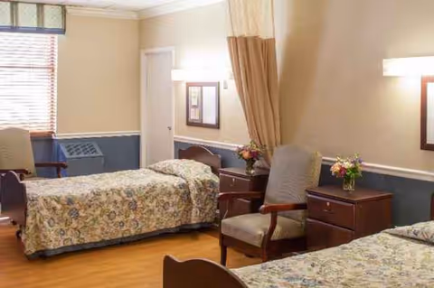 A shared senior living room with two beds, chairs, nightstands, and floral bedspreads in beige walls.