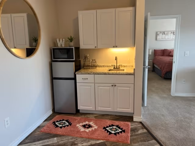 Small kitchenette area with white cabinets, a granite countertop with a sink, a microwave on top of a mini refrigerator, and a decorative pink rug with geometric patterns on the floor. A round mirror hangs on the adjacent wall. In the background, an open door reveals a bedroom with a bed covered in a red bedspread.