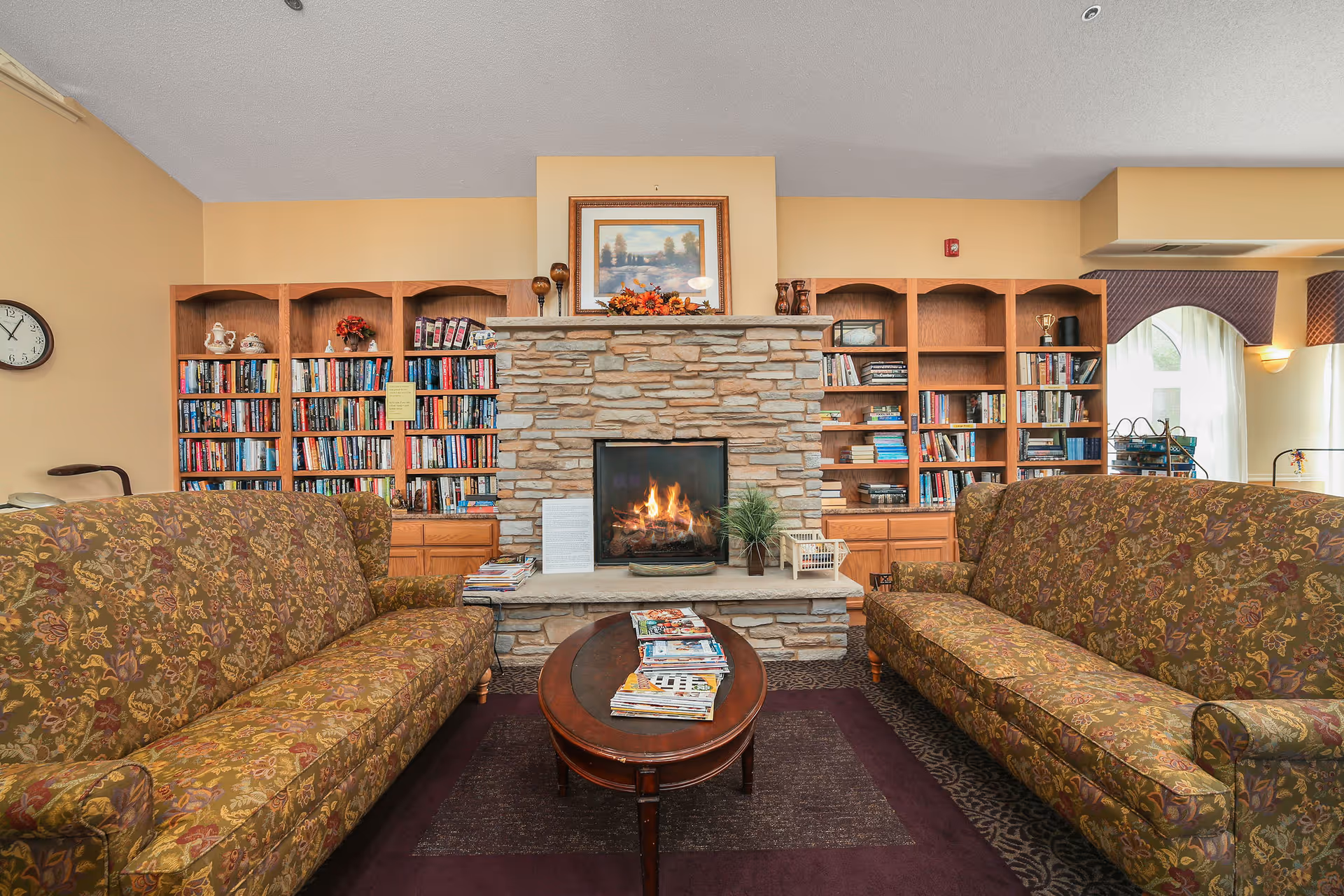 A cozy living room area with two patterned sofas facing each other across a wooden coffee table stacked with magazines. In the center is a stone fireplace with a fire burning, flanked by built-in wooden bookshelves filled with books and decorative items. Above the fireplace is a framed painting and some autumn-themed decorations. A wall clock is visible on the left wall.