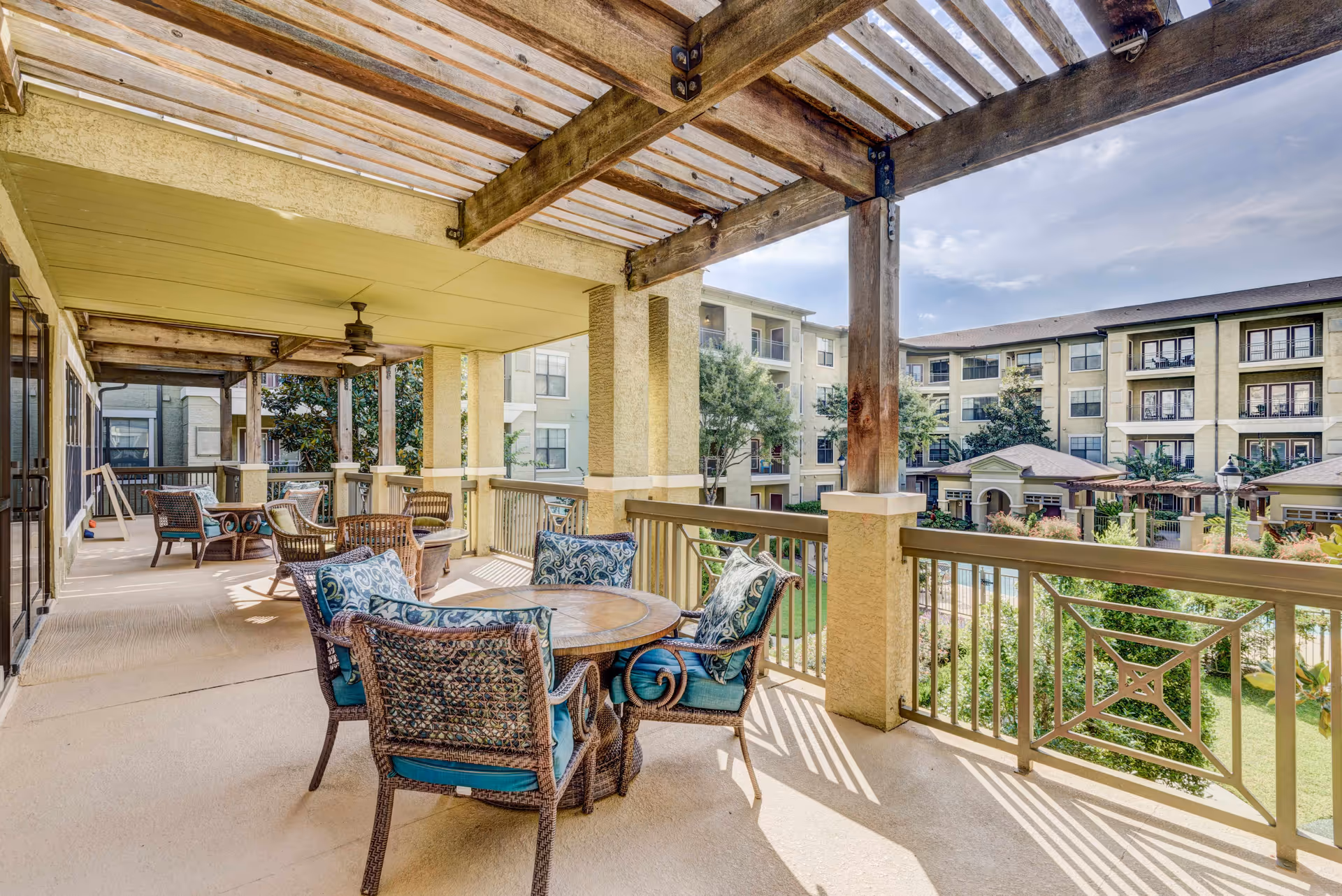 Covered outdoor patio with wicker chairs and tables overlooking a landscaped courtyard and surrounding apartment buildings.