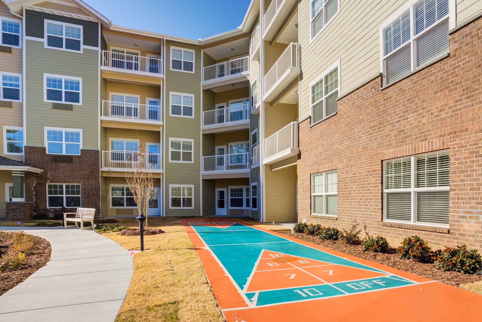 Outdoor courtyard area of a senior living facility with a shuffleboard court, a curved concrete walkway, a bench, and a multi-story building with balconies and windows under a clear blue sky.
