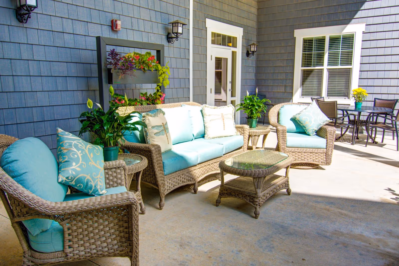 Outdoor patio area with wicker furniture including a sofa, two armchairs, and a glass-top coffee table. The cushions are light blue with decorative pillows featuring dragonfly designs. There are potted plants on side tables and a small dining table with chairs in the background. The area is adjacent to a building with gray siding, white trim around the door and window, and wall-mounted lantern-style lights.