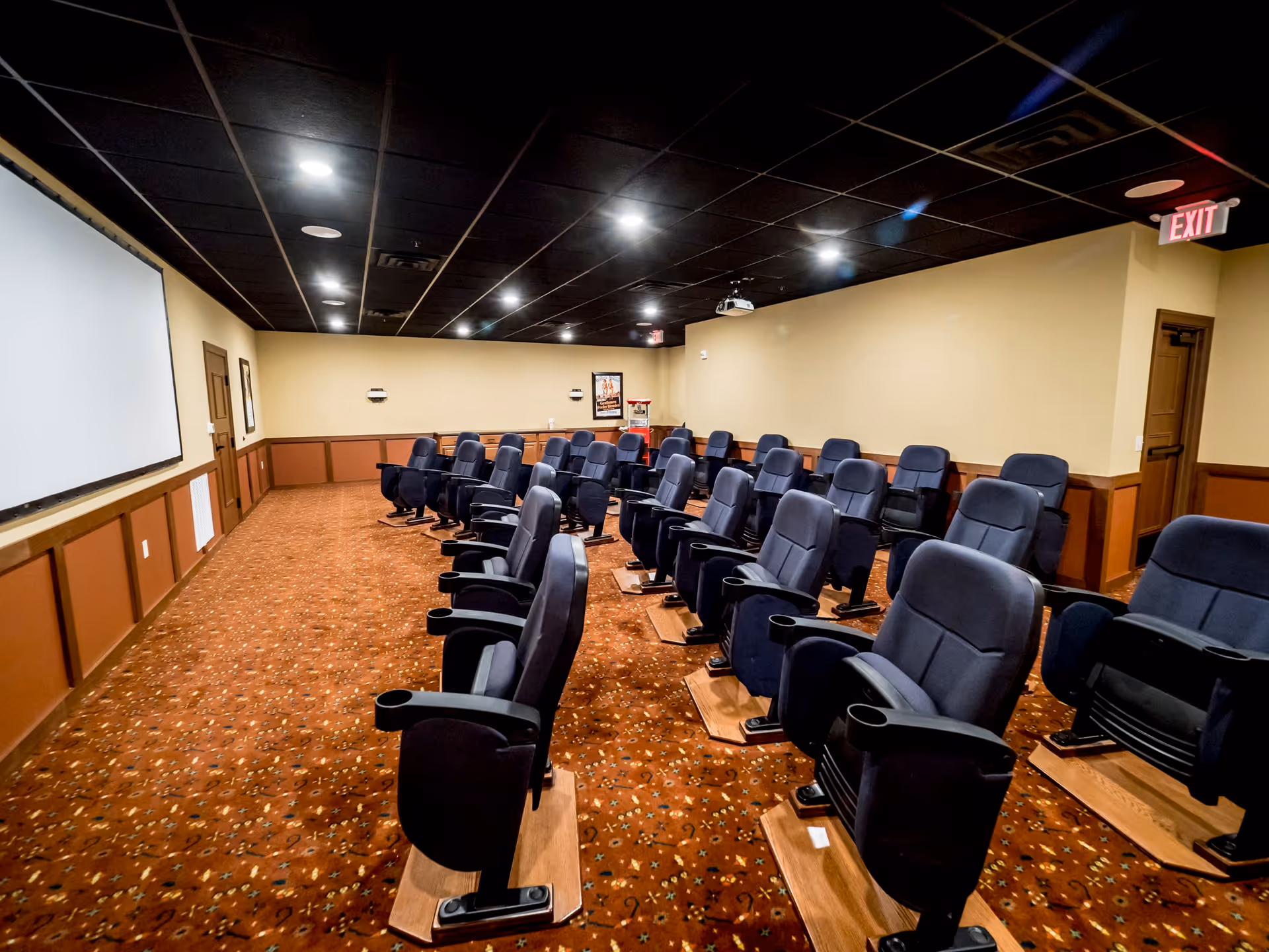 A small theater room with multiple rows of comfortable black reclining chairs with cup holders, facing a large white projection screen on the wall. The room has a patterned carpet, beige walls with wooden paneling, a black ceiling with recessed lighting, and an exit sign above a door in the back corner.