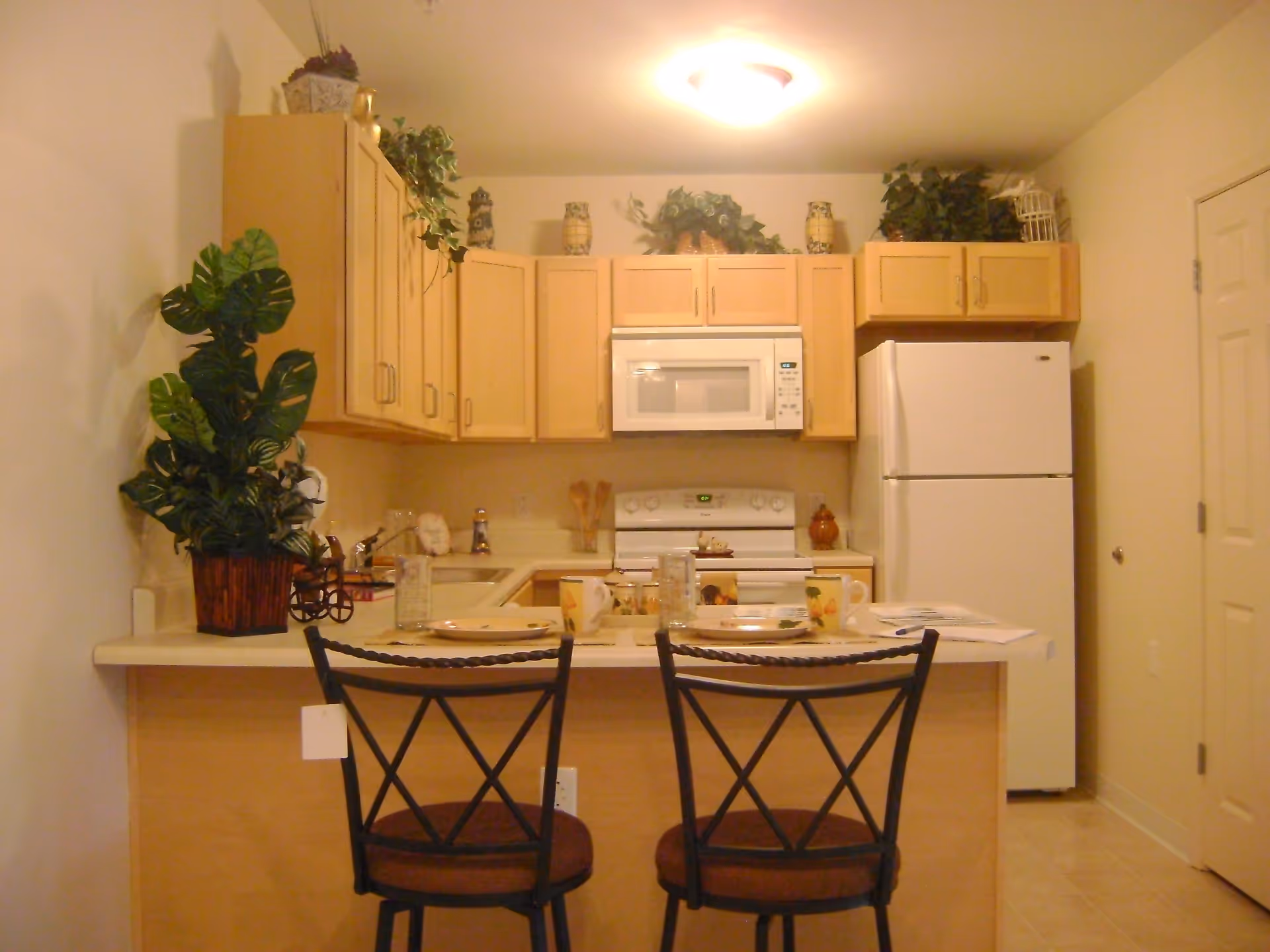 A small kitchen area with light wood cabinets, a white refrigerator, microwave, and stove. There is a countertop with two black metal chairs with brown cushions. The countertop is set with two plates, mugs, and glasses. Decorative plants and pottery are placed on top of the cabinets and countertop.