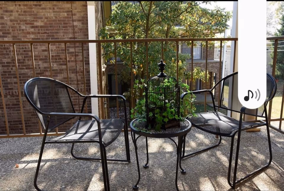 A small outdoor patio area with two black metal mesh chairs and a round black metal table. On the table is a decorative birdcage with green plants inside. The patio has a railing and overlooks a courtyard with trees and brick buildings.