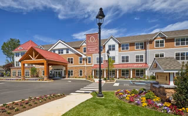 Exterior view of The Chelsea at Shrewsbury senior living facility showing a large three-story building with a red roof entrance canopy, well-maintained landscaping with colorful flowers, a lamppost with a sign that reads 'The Chelsea Welcome Home,' and a clear blue sky with some clouds.