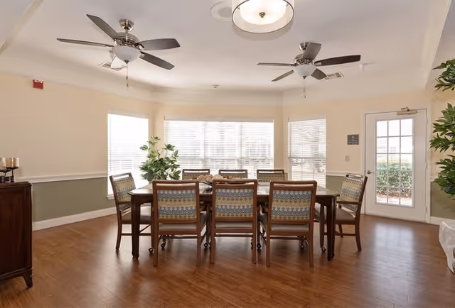 A bright dining room with a large rectangular wooden table surrounded by eight cushioned chairs with patterned upholstery. The room has wooden flooring, three large windows with white blinds, two ceiling fans with lights, a ceiling light fixture, a wooden cabinet on the left, and a glass door on the right leading outside. There are also green plants placed near the windows and door.