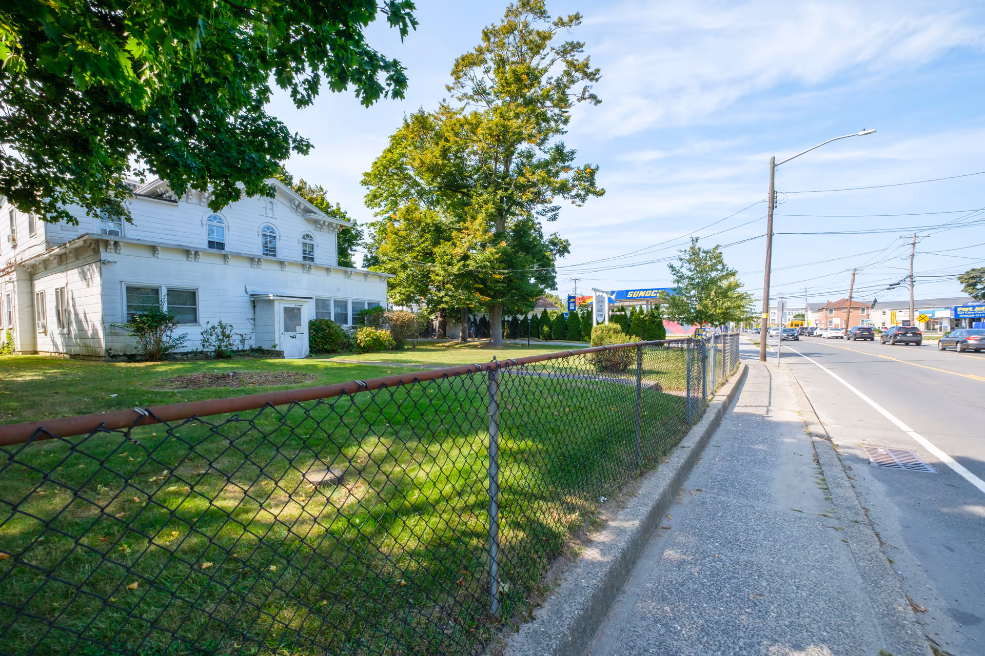 White two-story house set behind a chain-link fence with a tree-lined yard alongside a street and sidewalk.
