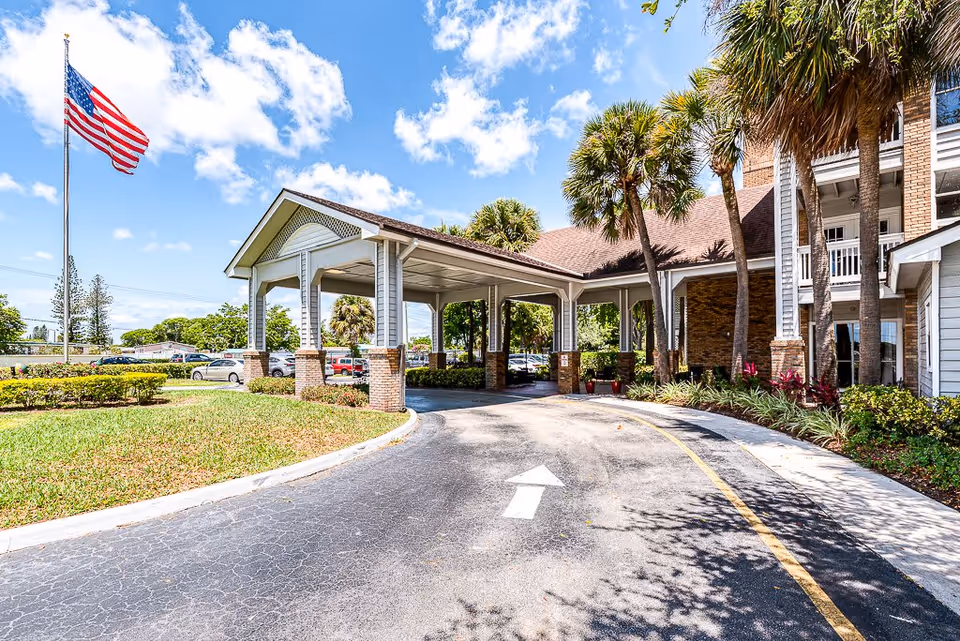 Exterior view of a senior living facility entrance with a covered drop-off area, palm trees, an American flag on a flagpole, and a driveway with an arrow pointing forward under a partly cloudy blue sky.