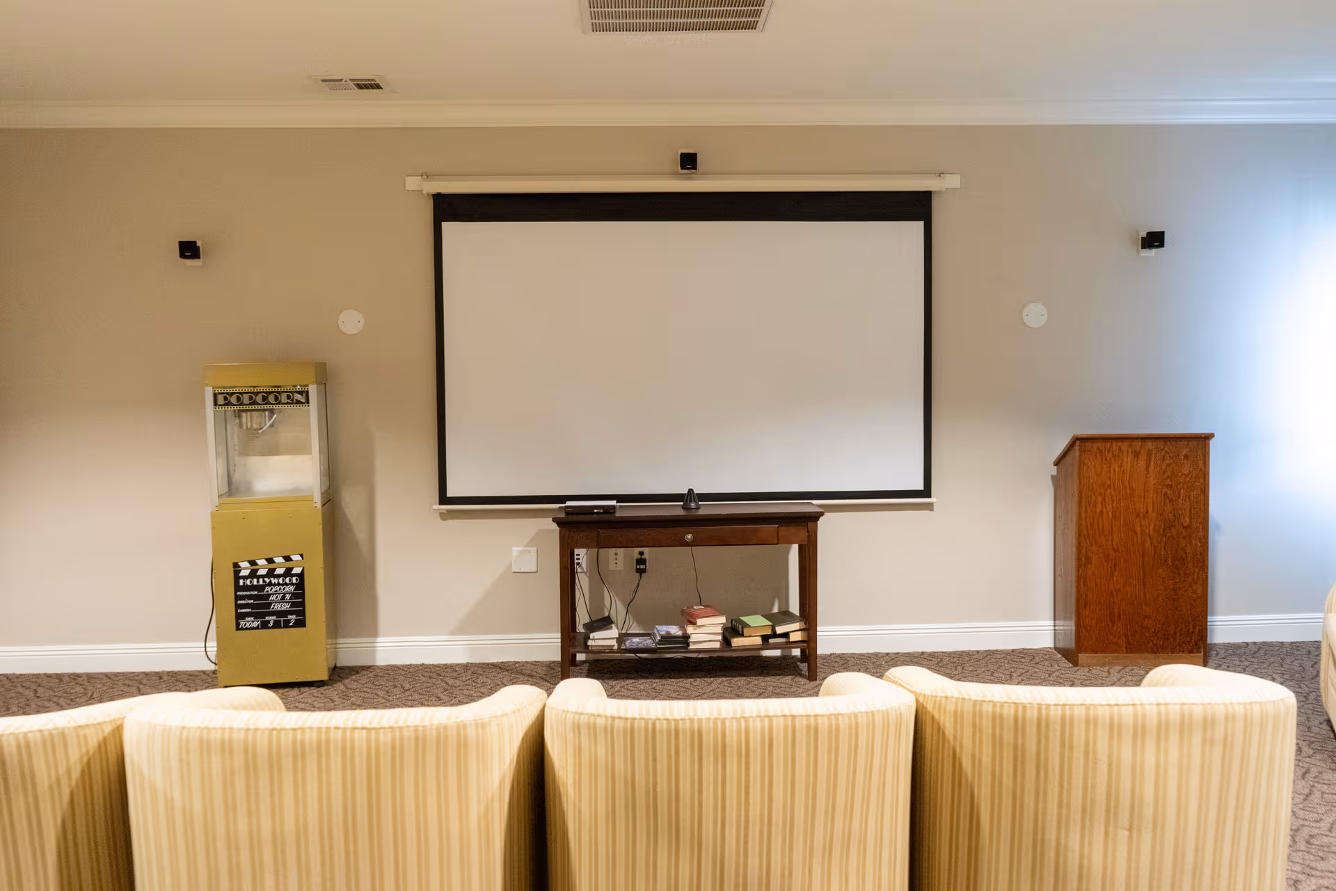 A small theater or presentation room with a large white projection screen mounted on a beige wall. In front of the screen is a wooden table holding books and electronic devices. To the left of the screen is a vintage-style popcorn machine, and to the right is a wooden podium. Four beige striped chairs are visible in the foreground facing the screen.