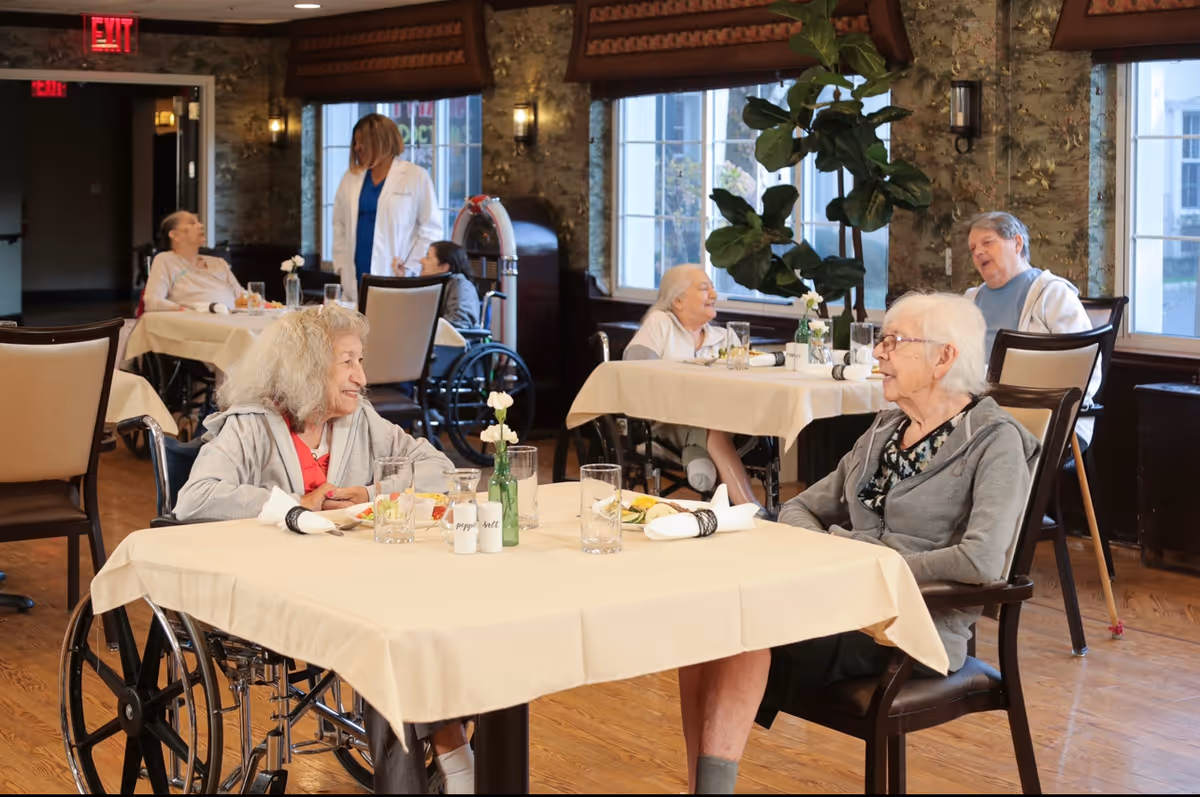 A dining room in a senior living facility with elderly residents seated at tables covered with beige tablecloths. Two women are seated at the foreground table, one in a wheelchair and the other in a chair, engaged in conversation. In the background, other residents and a staff member in a white coat are visible. The room has large windows, wooden floors, and decorative wall sconces.