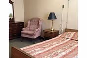 A bedroom with a wooden bed covered with a patterned bedspread, a wooden nightstand with a brass lamp, a pink upholstered armchair, and a wooden dresser with a mirror.
