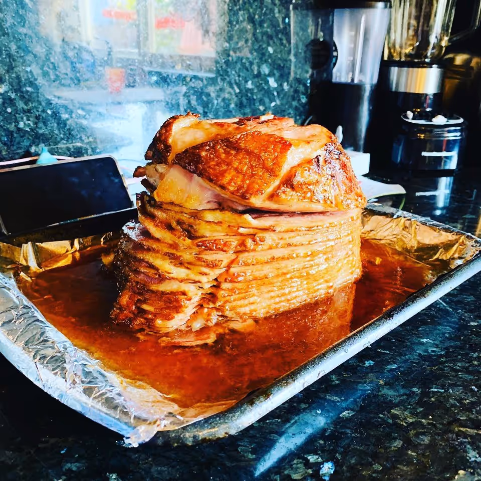 A cooked spiral ham placed on a foil-lined baking tray with juices around it, set on a dark granite kitchen countertop with kitchen appliances in the background.