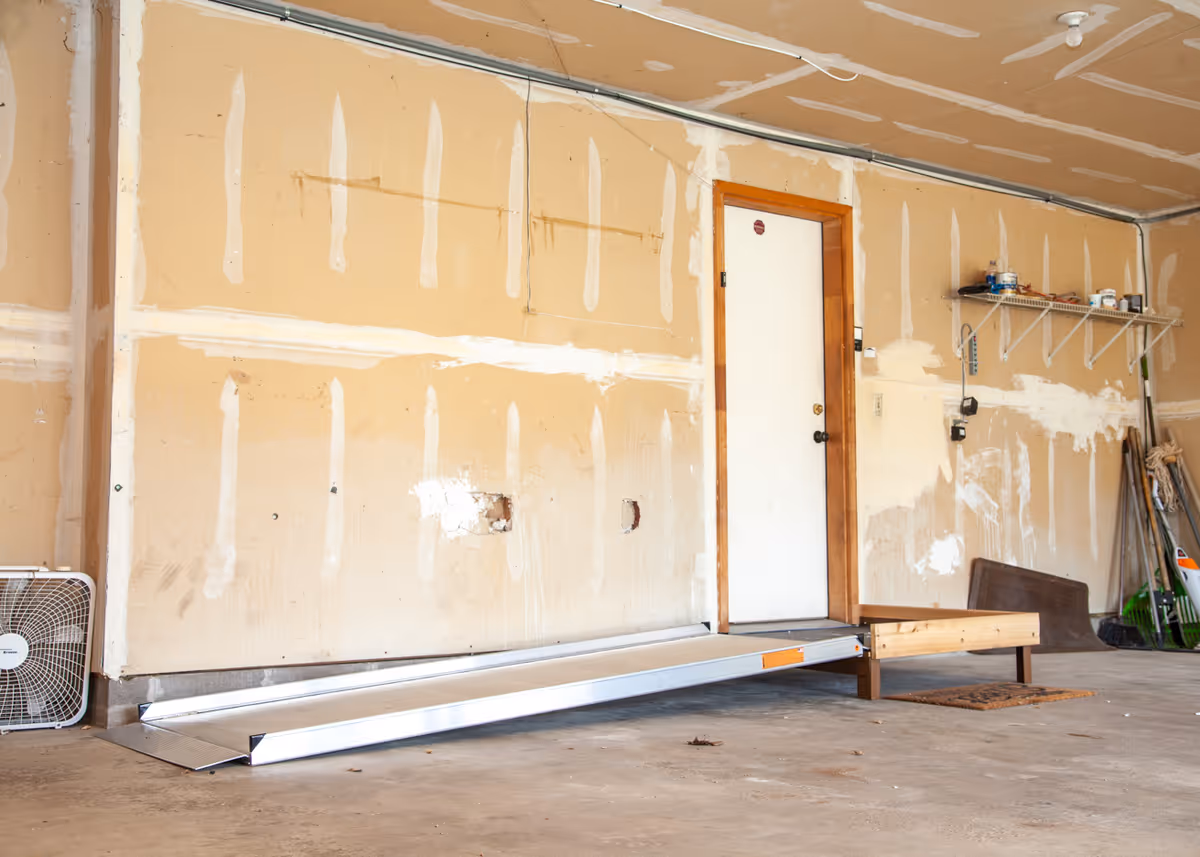 Unfinished garage interior with a metal access ramp leading to a door, exposed drywall, shelving and tools.