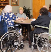 Four elderly individuals, two in wheelchairs, sitting around a round table playing cards in a cozy room with wooden furniture and a framed picture on the wall.