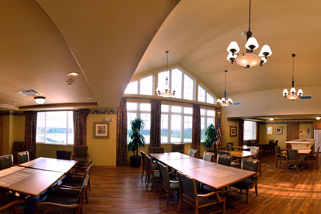 Spacious dining room with long wooden tables and chairs, large arched windows, plants, and hanging chandeliers.